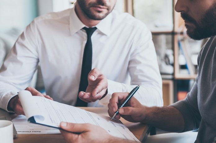 Signing contract. Close-up of confident young man signing some document while another man in shirt and tie sitting close to him and pointing document